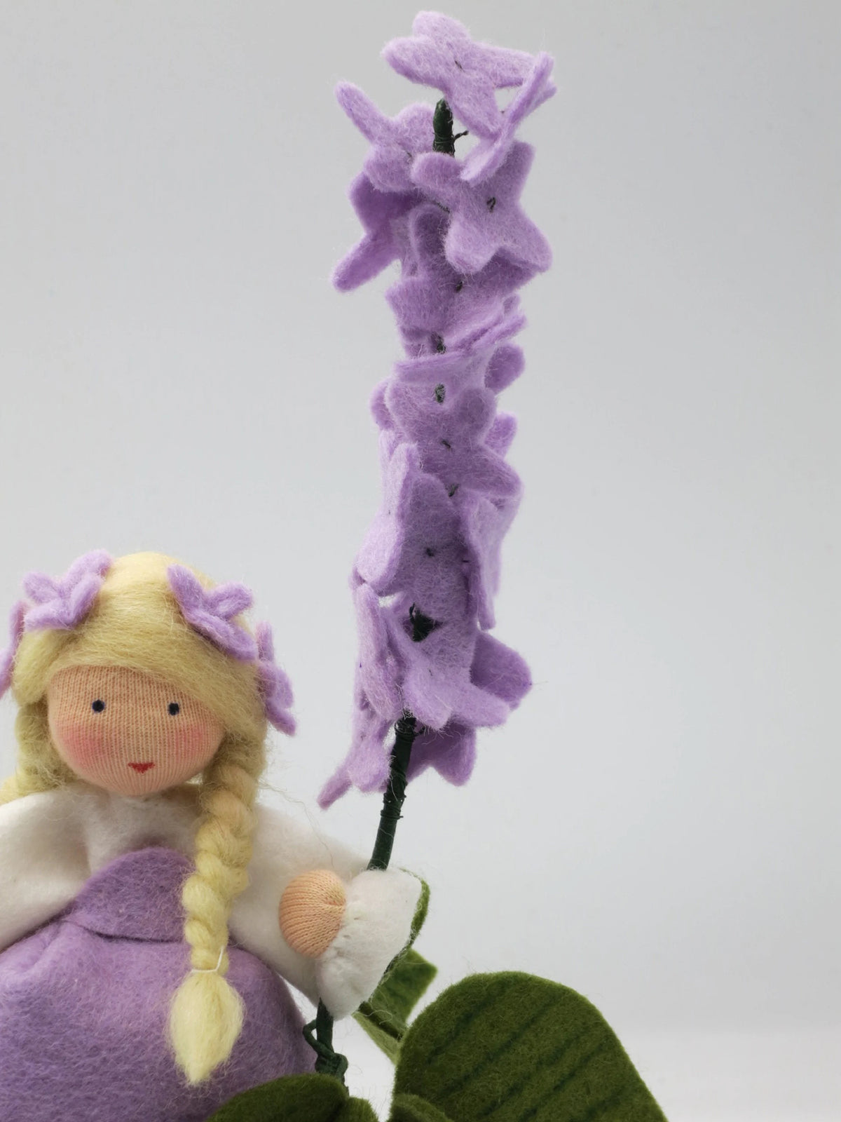 Detailansicht des handgefertigten Flieder-Blumenkinds aus Wollfilz mit lila Fliederblüten, blondem Zopf und grünem Blatt.