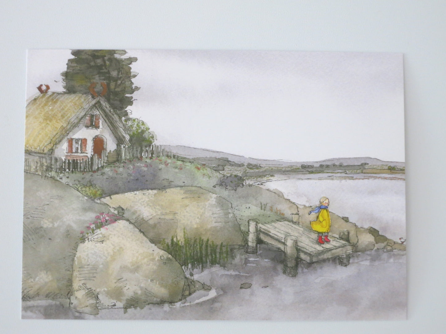 Kind im gelben Regenmantel auf einem Steg vor reetgedecktem Haus am See – Postkarte Mädchen am See von Sanne Dufft
