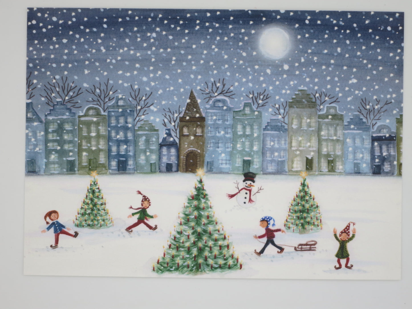 Winterpostkarte für Kinder: Kinder beim Schlittschuhlaufen vor verschneiten Giebelhäusern mit Weihnachtsbäumen und Vollmond