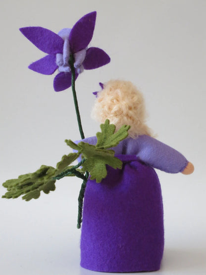 Rückansicht des Akelei-Blumenkinds mit violettem Filzkleid, großer Blüte und grünem Blattzweig.