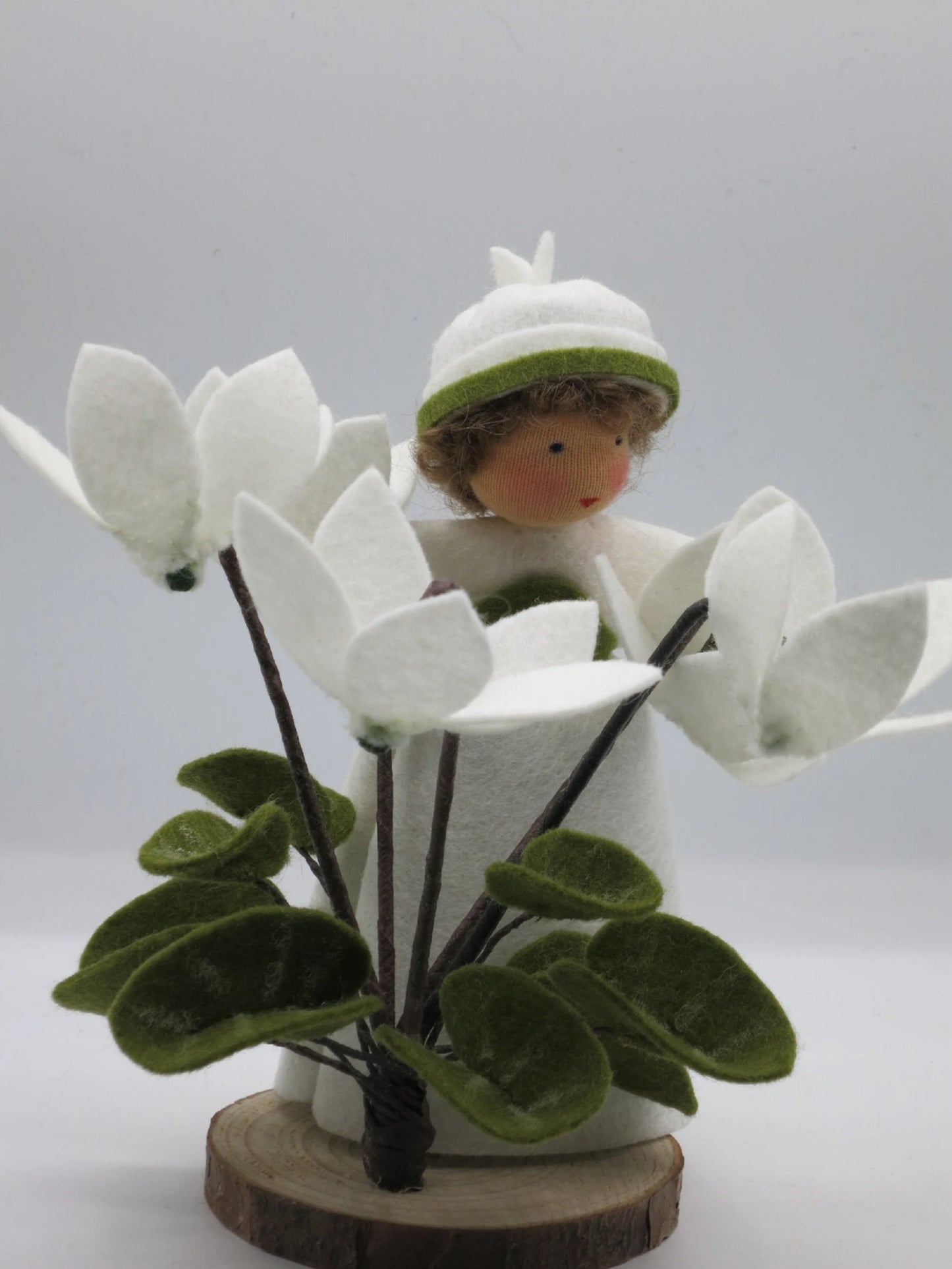 Handgefertigtes Alpenveilchen-Blumenkind mit weißen Filzblüten und grünen Blättern auf einer Holzscheibe.