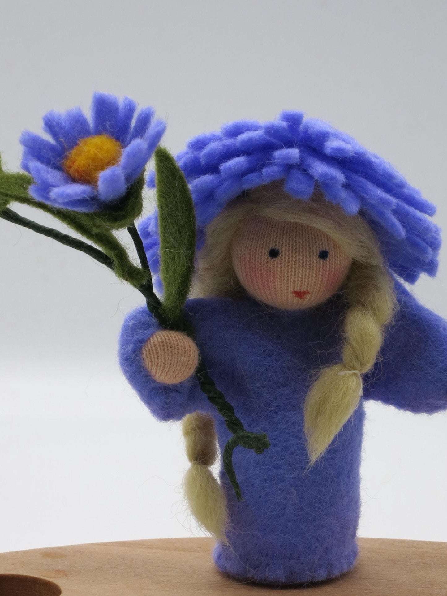 Aster-Blumenkind Art. HE54 mit violetter Blütenhaube, blondem Zopf und Asternblüte auf dem Geburtstagsring.