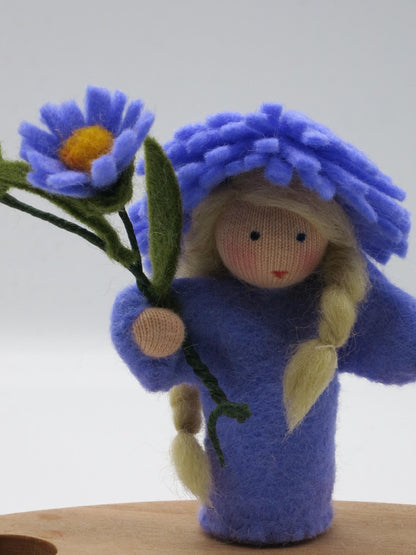 Aster-Blumenkind Art. HE54 mit violetter Blütenhaube, blondem Zopf und Asternblüte auf dem Geburtstagsring.