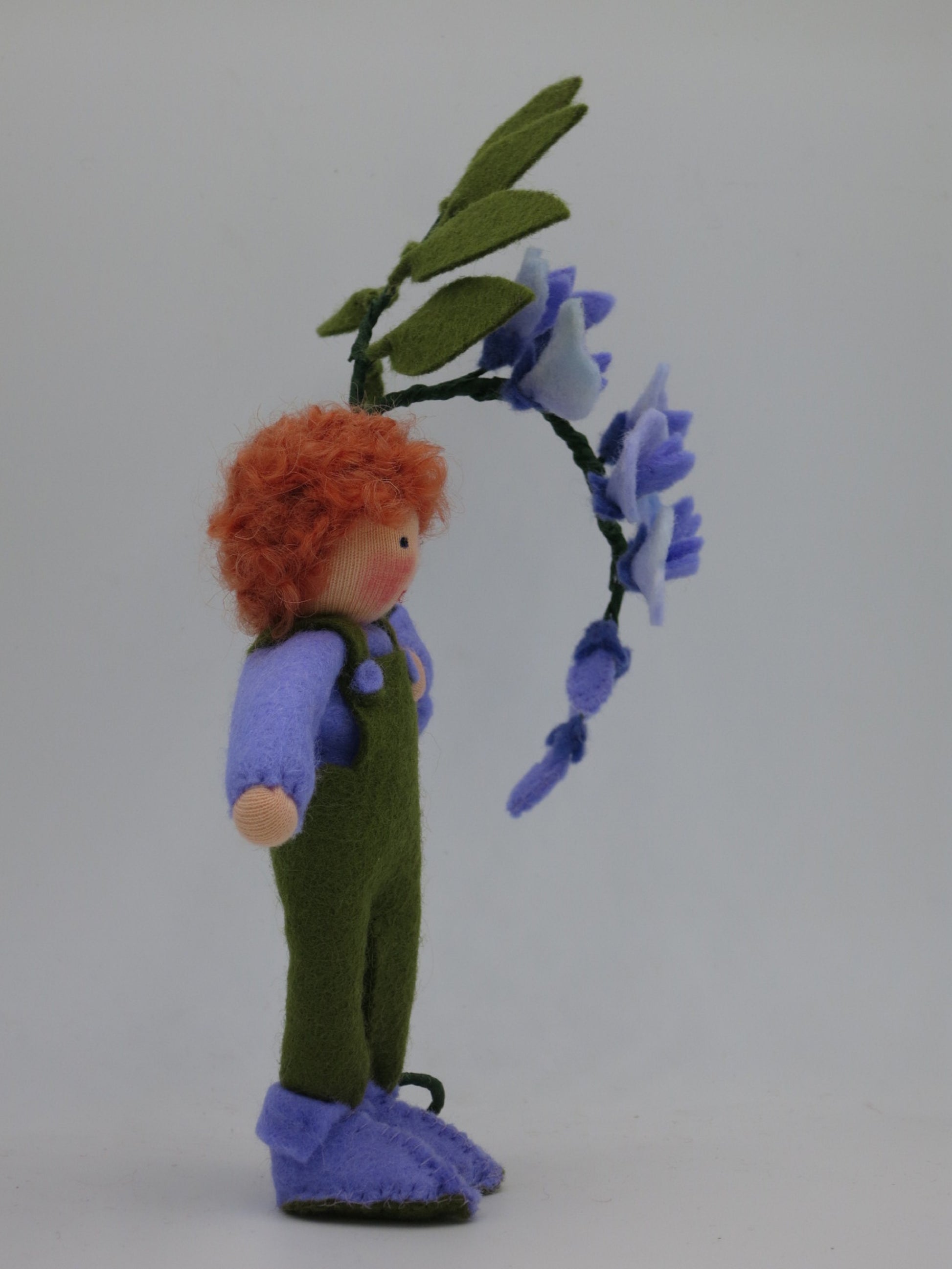 Profilansicht des handgefertigten Blauregen-Blumenkinds mit rotem Wollhaar, grüner Filzhose und violettem Blütenzweig.