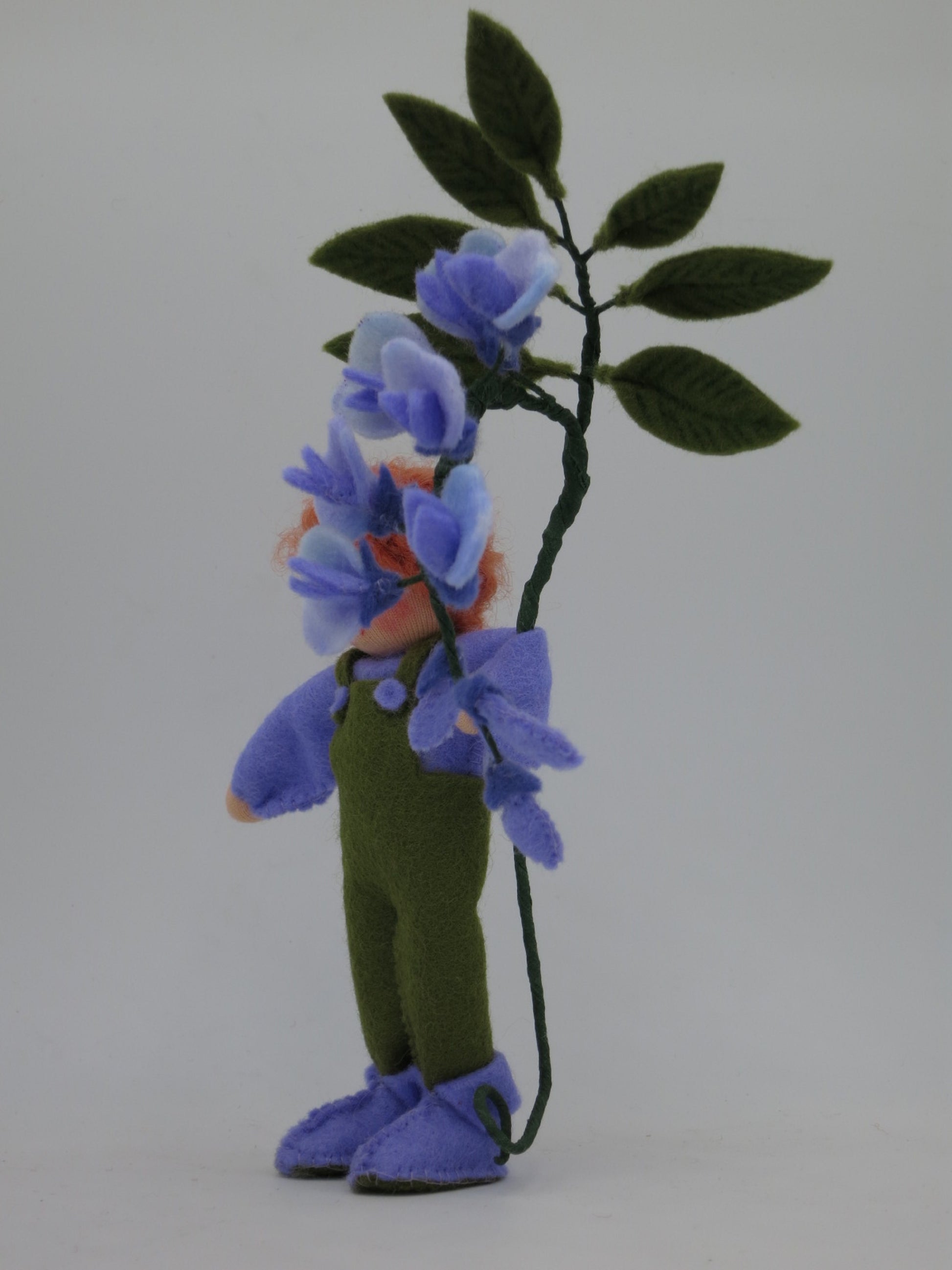 Seitenansicht des Blauregen-Blumenkinds in grün-violetter Filzkleidung, umrahmt von herabhängenden Blütentrauben.