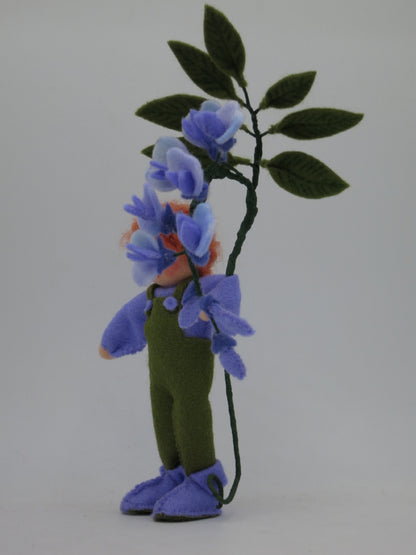 Seitenansicht des Blauregen-Blumenkinds in grün-violetter Filzkleidung, umrahmt von herabhängenden Blütentrauben.