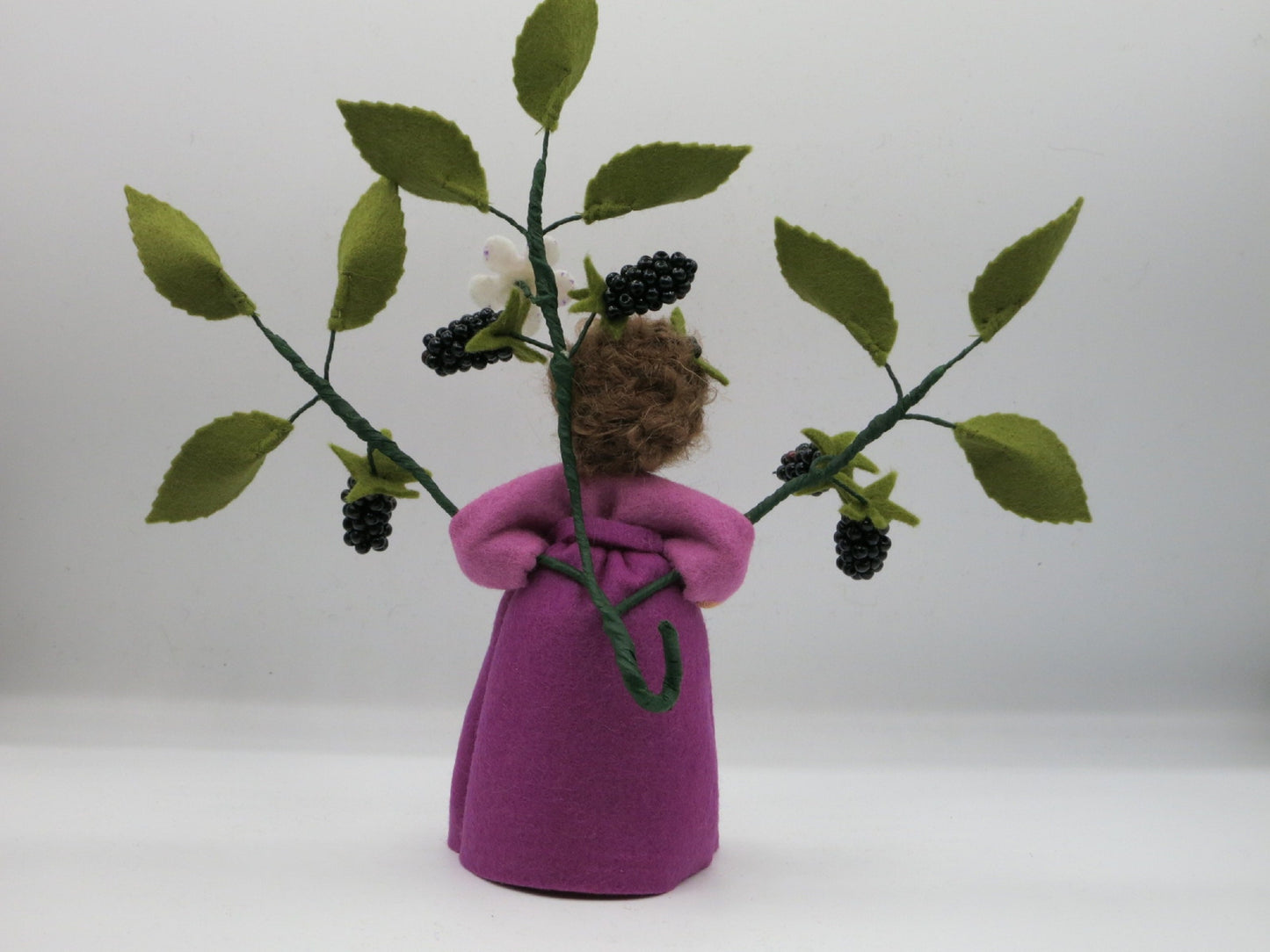 Rückansicht des Brombeer-Blumenkinds mit braunem Wollhaar und ausgebreiteten Brombeerzweigen mit grünen Blättern.