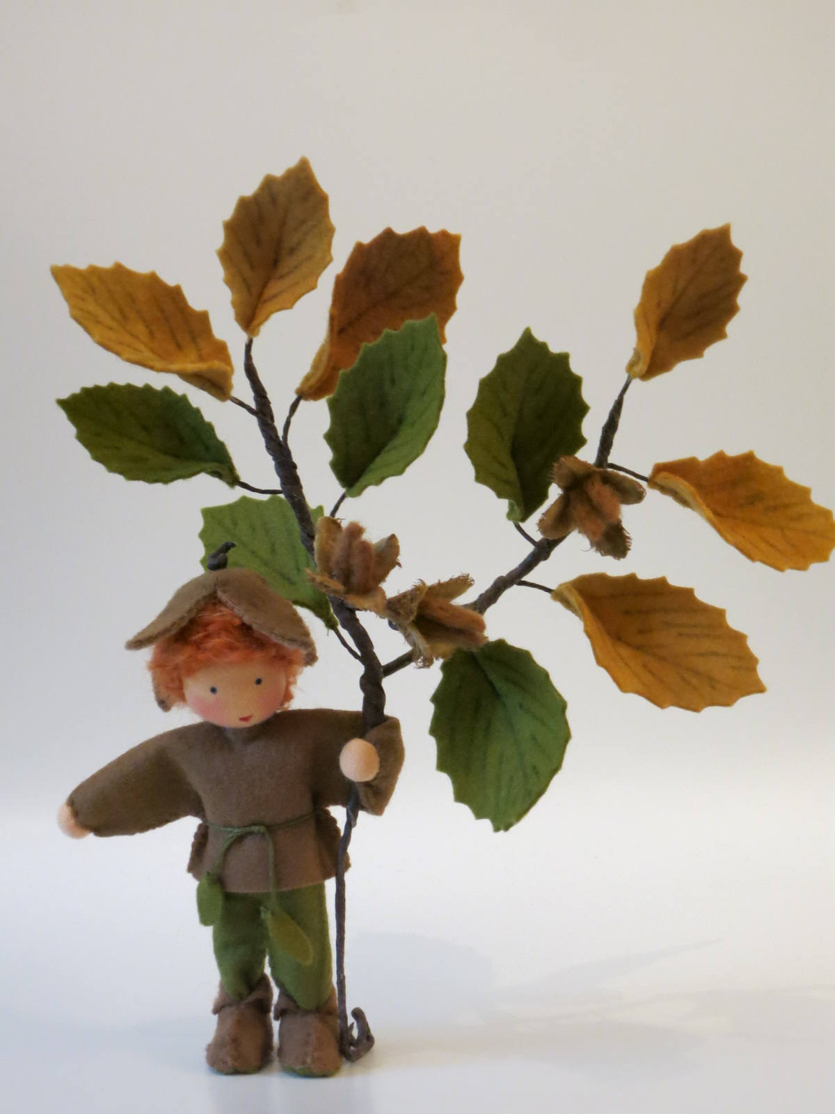 Gesamtansicht des Bucheckern-Jungen mit braunem Filzhut, grünem Beinkleid und hohem Buchenzweig in Herbstfarben.