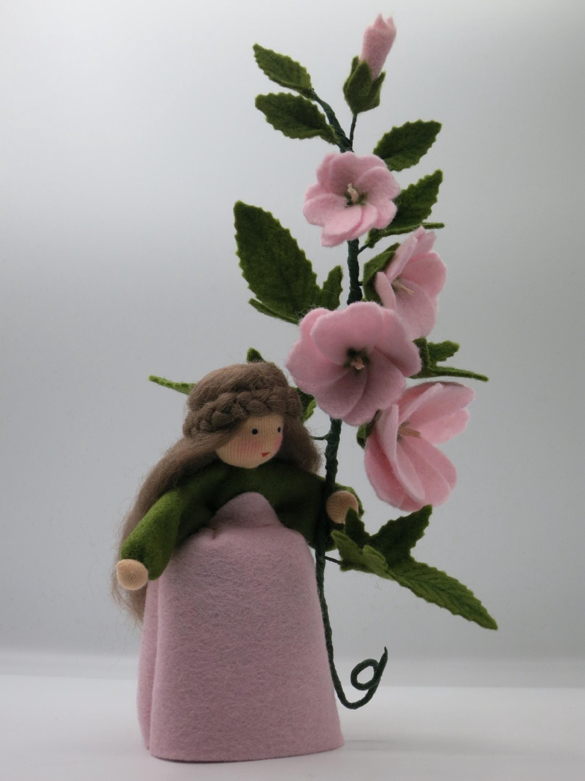 Buschmalve-Blumenkind in Frontalansicht mit rosa Filzkleid, grüner Stola und hohem Blütenzweig für den Sommer