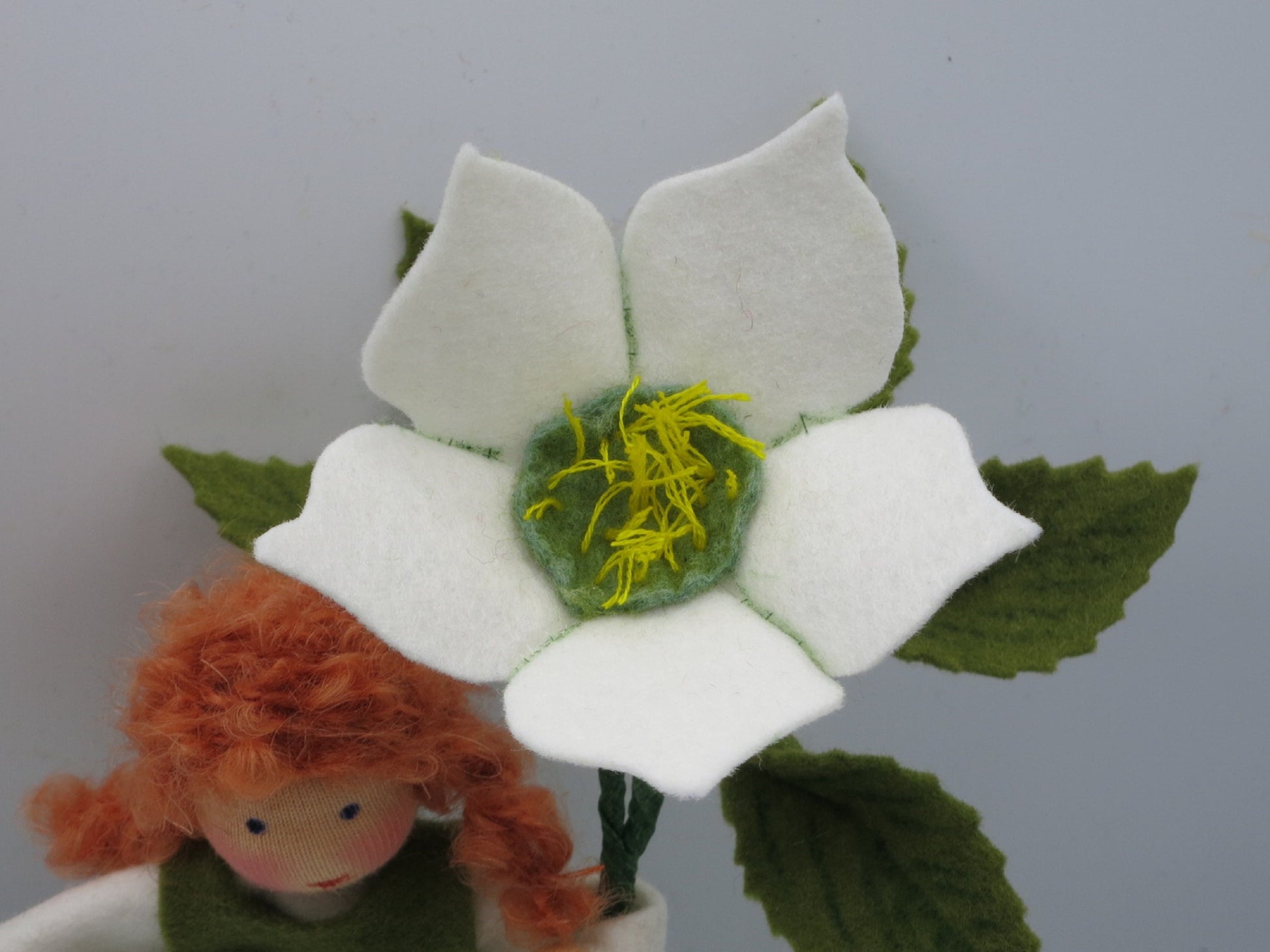 Detail einer weißen Christrose aus Filz mit grün-gelber Mitte, Blättern und rothaarigem Blumenkind