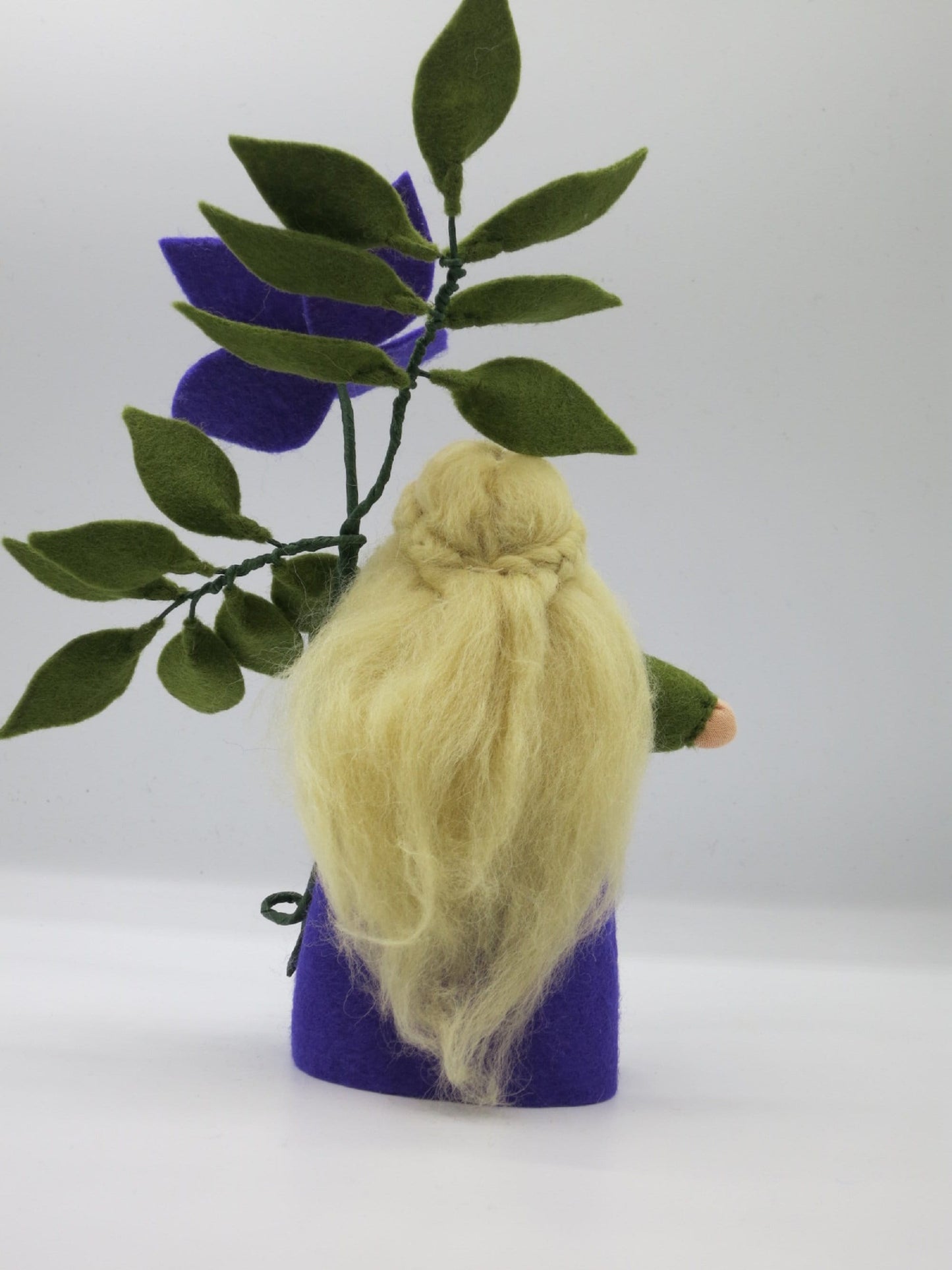 Rückansicht des Clematis-Blumenkinds mit langem blondem Wollhaar, violettem Filzkleid und grünem Blätterzweig