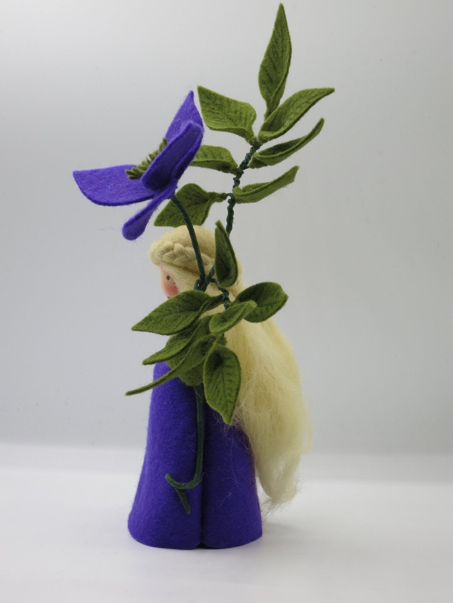 Seitliche Ansicht des Clematis-Blumenkinds mit langem blondem Wollhaar, violettem Filzkleid und Blätterzweig