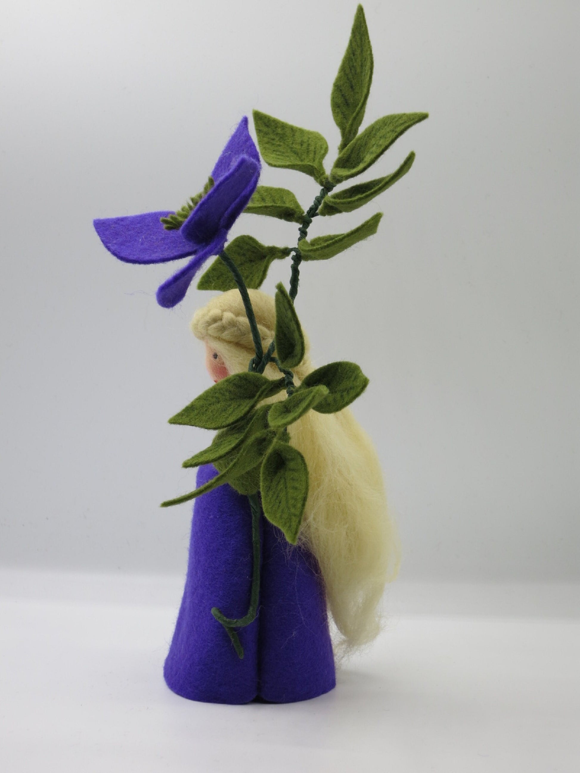 Seitliche Ansicht des Clematis-Blumenkinds mit langem blondem Wollhaar, violettem Filzkleid und Blätterzweig