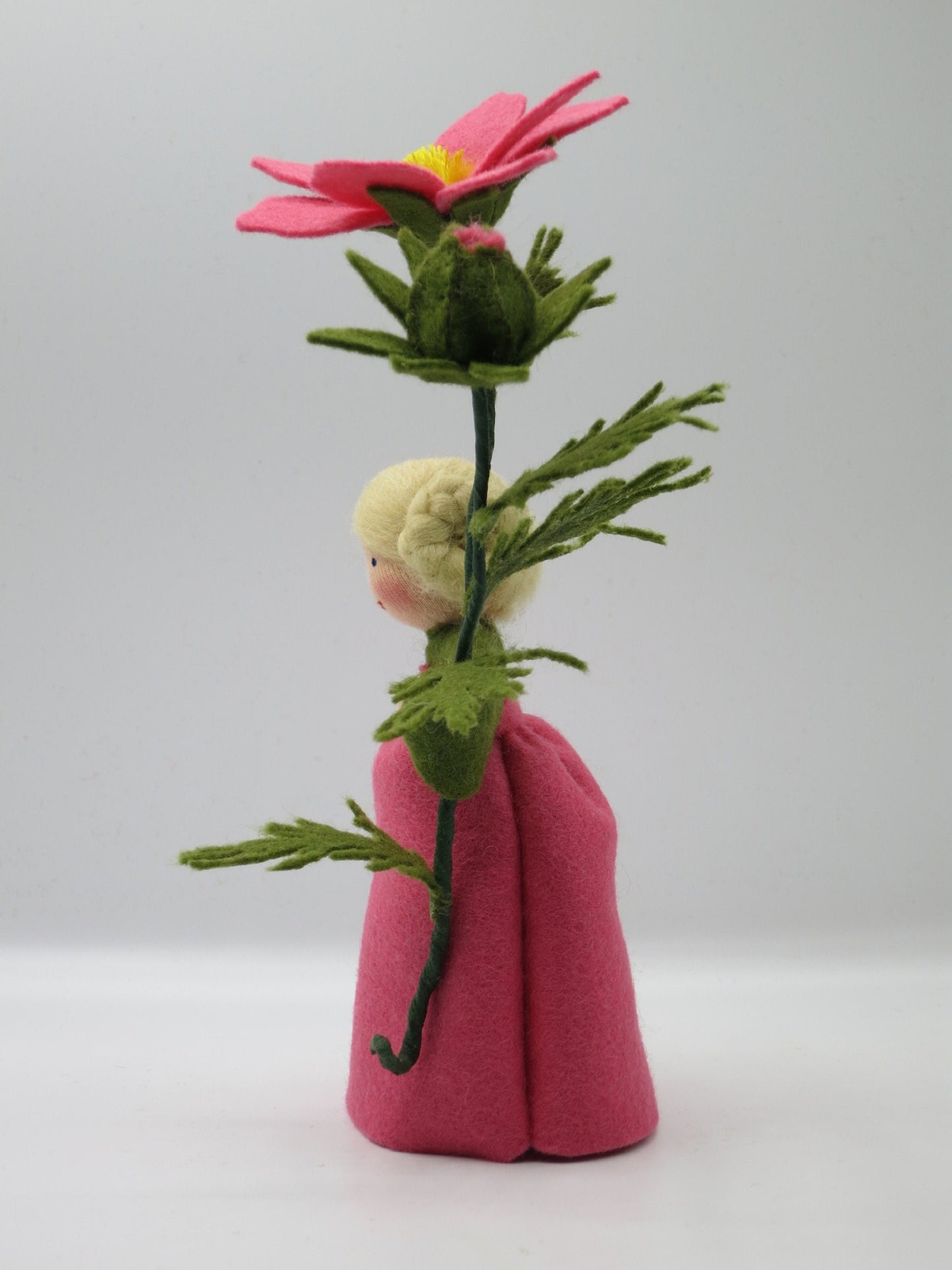 Seitliche Ansicht des Cosmea-Blumenkinds im rosa Filzkleid mit hoher Blüte, Knospe und feinem grünem Blattwerk