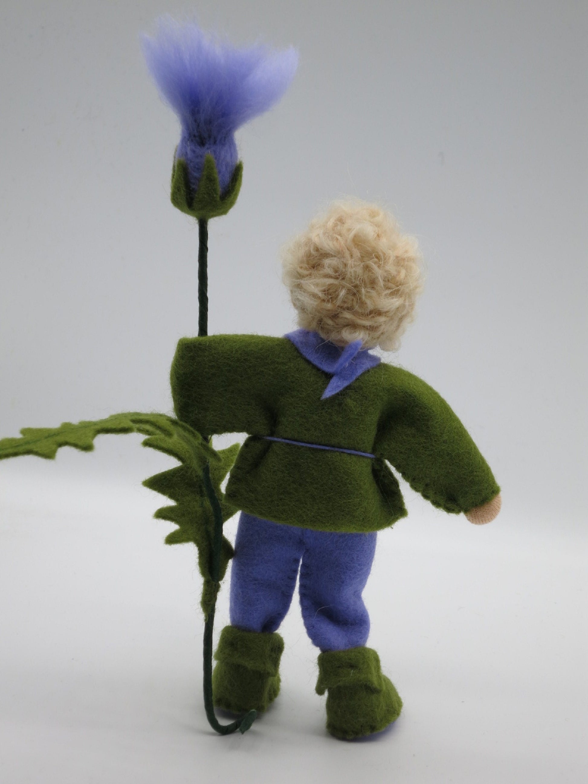 Rückansicht des Distel-Jungen mit blondem Lockenhaar, grüner Filzjacke, lila Hose und hoher Distelblüte