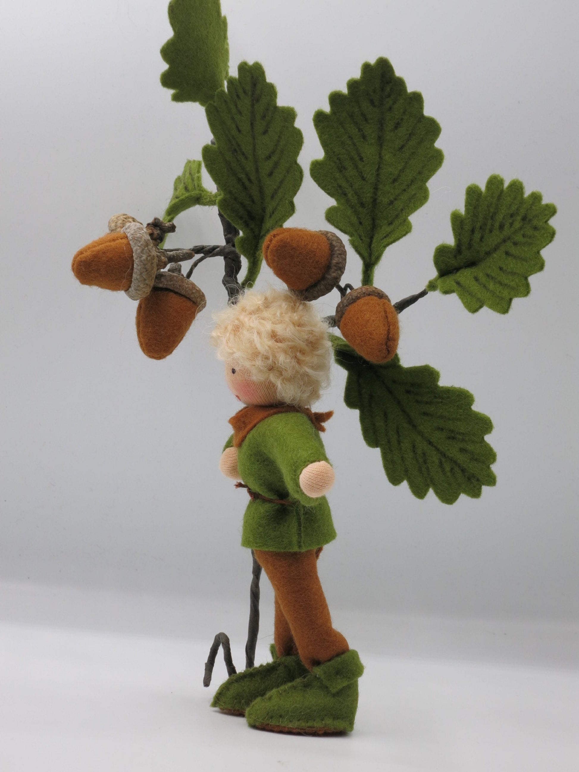 Seitenansicht des Eichel-Jungen aus Filz vor einem Eichenzweig mit Eicheln als Waldorf-Herbstdeko.