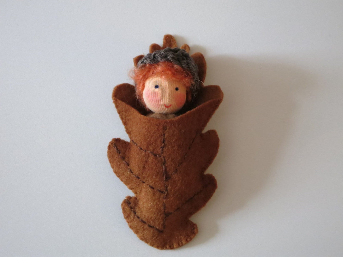 Eichelbaby mit rotem Haar als handgefertigte Filzpuppe in einer braunen Blätterhülle für die Herbstdekoration.