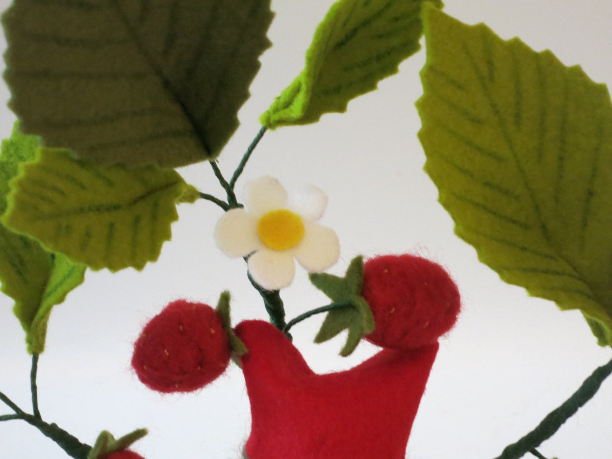 Detailansicht des Erdbeer-Blumenkinds aus Wollfilz mit weißen Erdbeerblüten, roten Früchten und grünen Blättern.