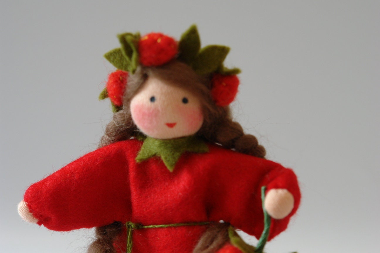 Detailansicht des Erdbeer-Blumenkinds aus Wollfilz mit roten Erdbeeren im Haar, rotem Kleid und grünen Blättern.