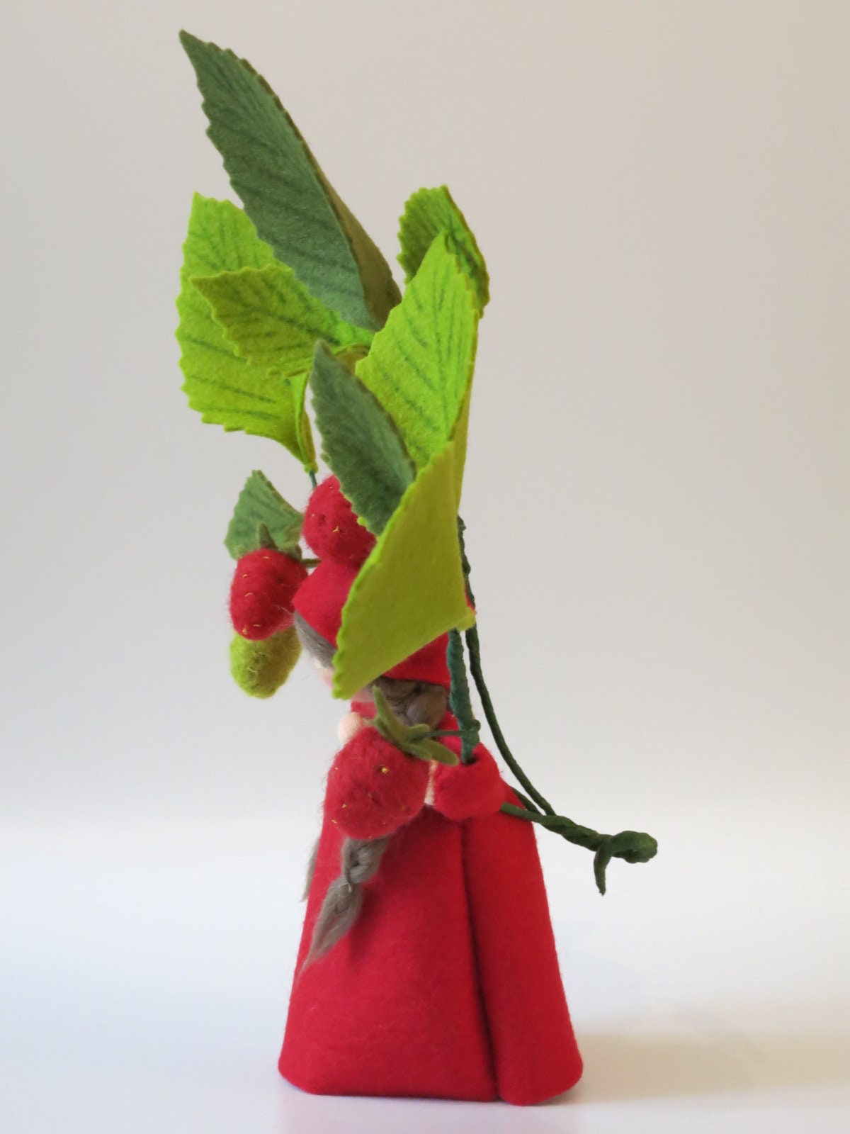 Seitenansicht des Erdbeer-Blumenkinds aus Wollfilz mit rotem Kleid, reifen Erdbeeren und großem grünem Blattzweig.
