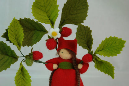 Erdbeer-Blumenkind aus Wollfilz unter einem Zweig mit roten Erdbeeren und grünen Blättern als Sommerdekoration.