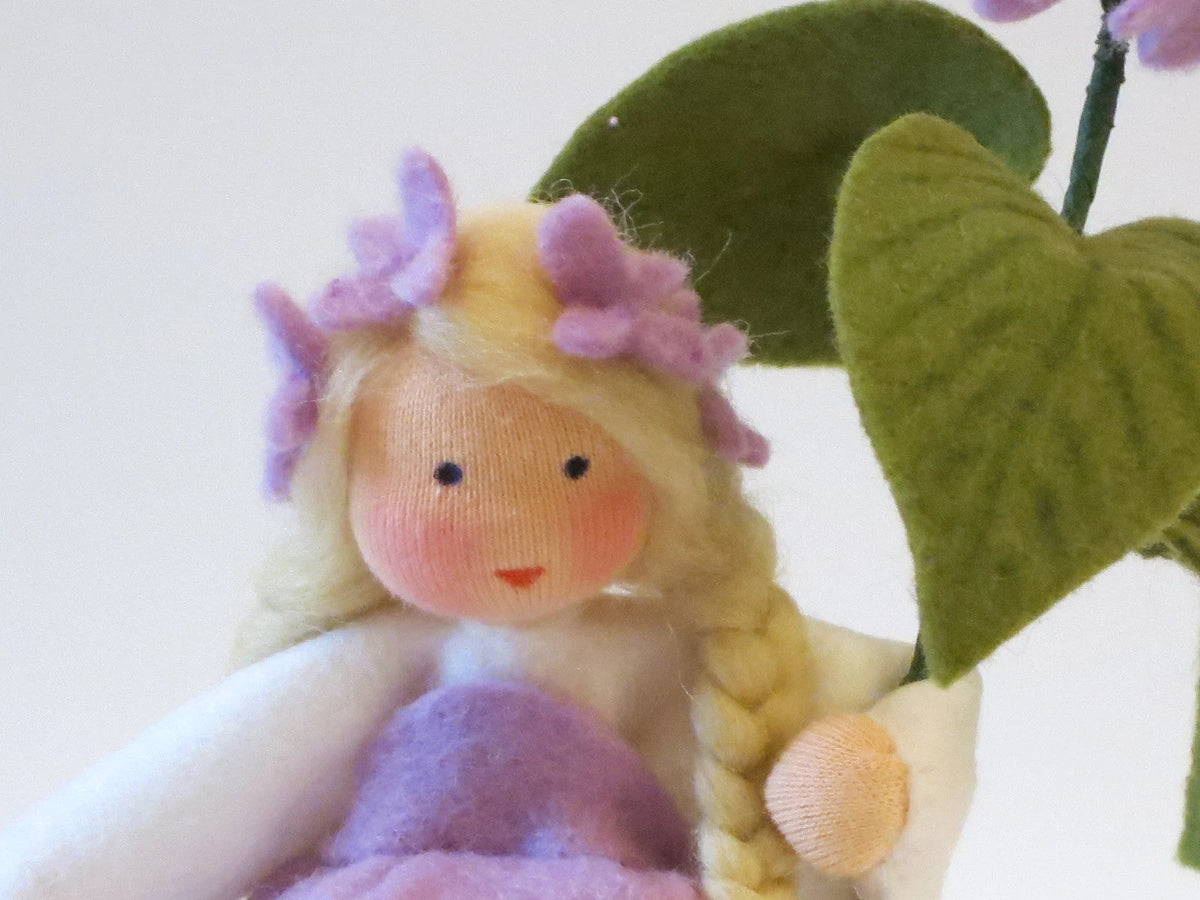 Porträt des Flieder-Blumenkinds aus Wollfilz mit blondem Zopf, lila Fliederblüten und großen grünen Blättern.