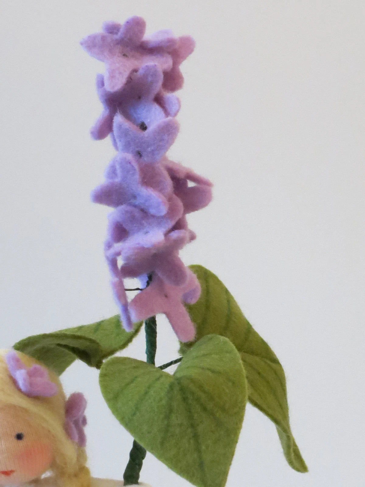 Detailansicht des Flieder-Blumenkinds aus Wollfilz mit lila Fliederblüten und großen grünen Blättern.