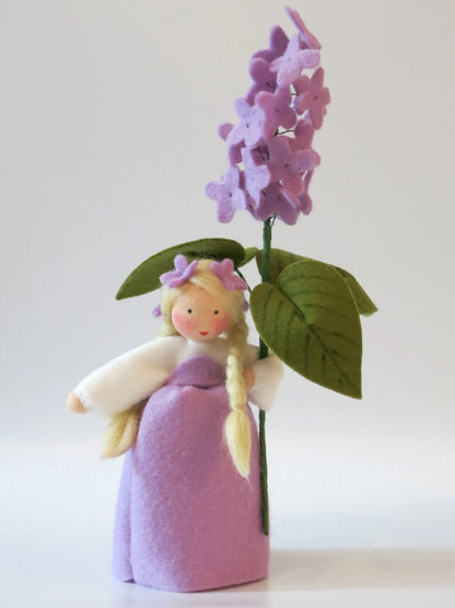 Frontalansicht des handgefertigten Flieder-Blumenkinds aus Wollfilz mit lila Kleid, Zöpfen und blühendem Fliederzweig.