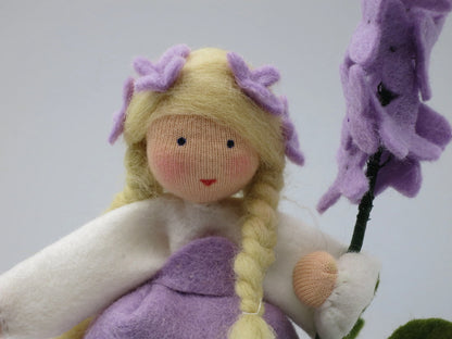 Nahaufnahme des Flieder-Blumenkinds aus Wollfilz mit lila Kleid, blondem Zopf und Fliederblütenzweig.