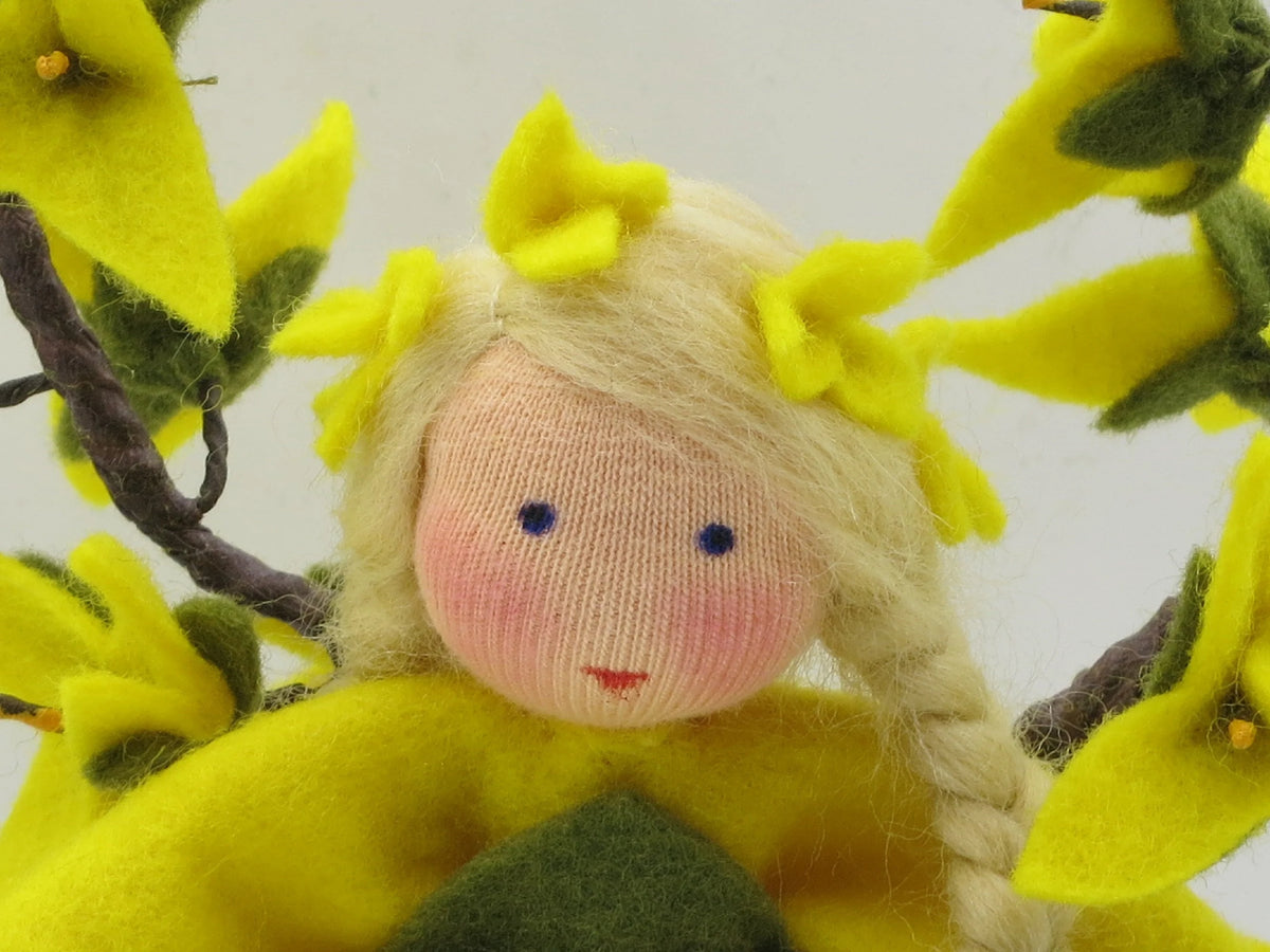 Nahaufnahme des Forsythien-Blumenkinds aus Wollfilz mit blondem Haar und gelben Forsythienblüten.