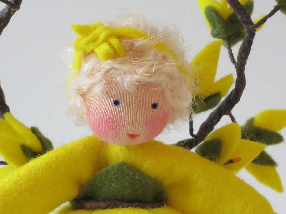 Porträt des Forsythien-Blumenkinds aus Wollfilz mit blondem Haar, gelben Blüten im Haar und Zweigen im Hintergrund.