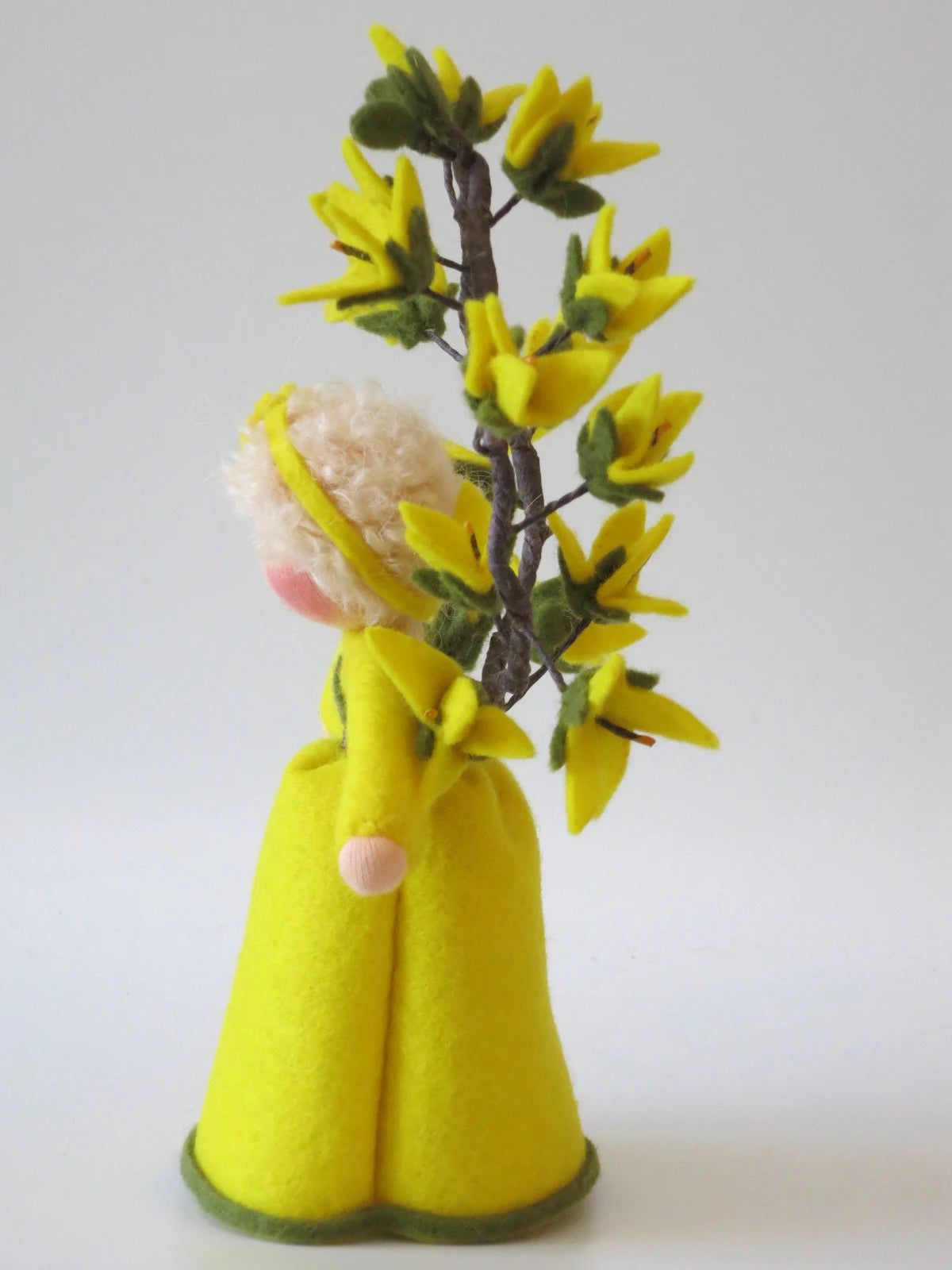 Seitenansicht des Forsythien-Blumenkinds aus Wollfilz mit gelbem Kleid und hoch aufragendem Zweig voller gelber Blüten.