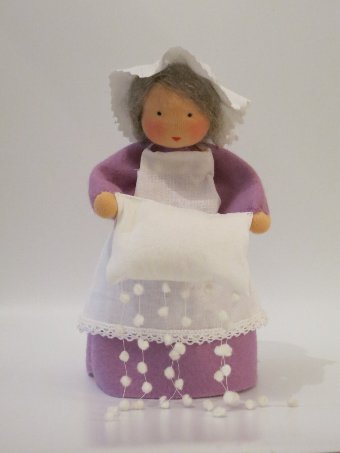 Handgefertigte Frau Holle aus Wollfilz mit lila Kleid, weißer Schürze und Kissen, aus dem Schneeflocken fallen.