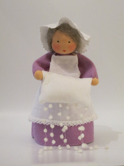 Handgefertigte Frau Holle aus Wollfilz mit lila Kleid, weißer Schürze und Kissen, aus dem Schneeflocken fallen.