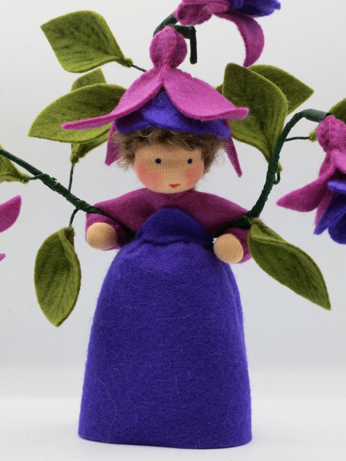 Frontalansicht des Fuchsien-Blumenkinds aus Wollfilz mit violettem Kleid, pink-violettem Blütenhut und Fuchsienzweigen.
