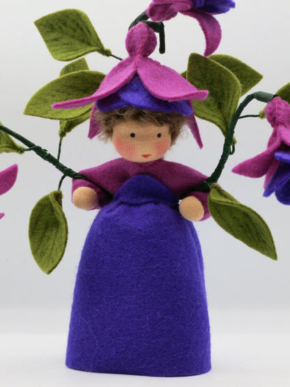 Frontalansicht des Fuchsien-Blumenkinds aus Wollfilz mit violettem Kleid, pink-violettem Blütenhut und Fuchsienzweigen.