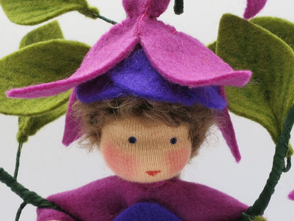 Nahaufnahme des Fuchsien-Blumenkinds aus Wollfilz mit braunem Haar, violettem Blütenhut und grünen Blättern.