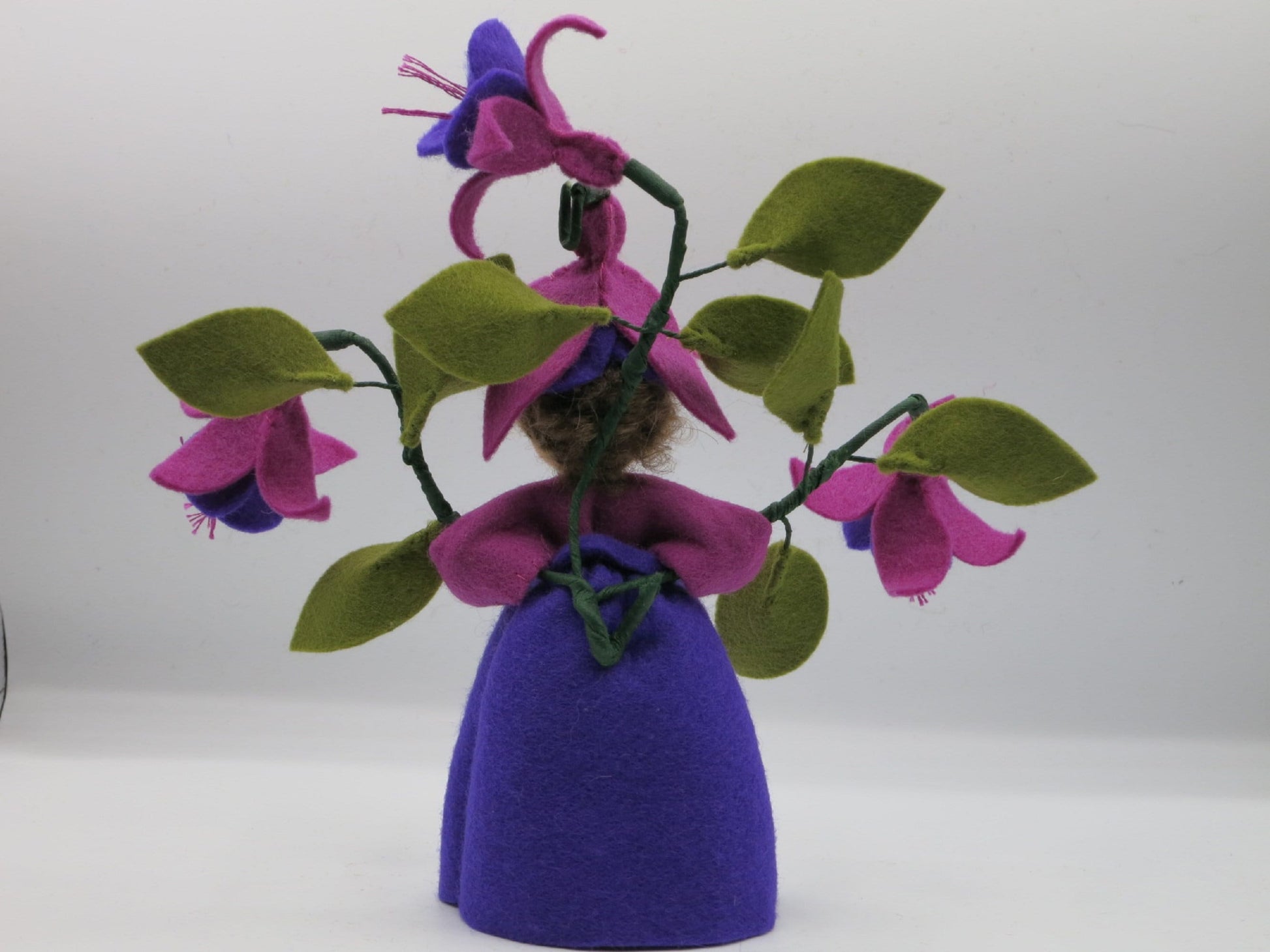 Hinteransicht des Fuchsien-Blumenkinds aus Wollfilz mit violettem Kleid, grünen Blättern und hängenden Fuchsienblüten.