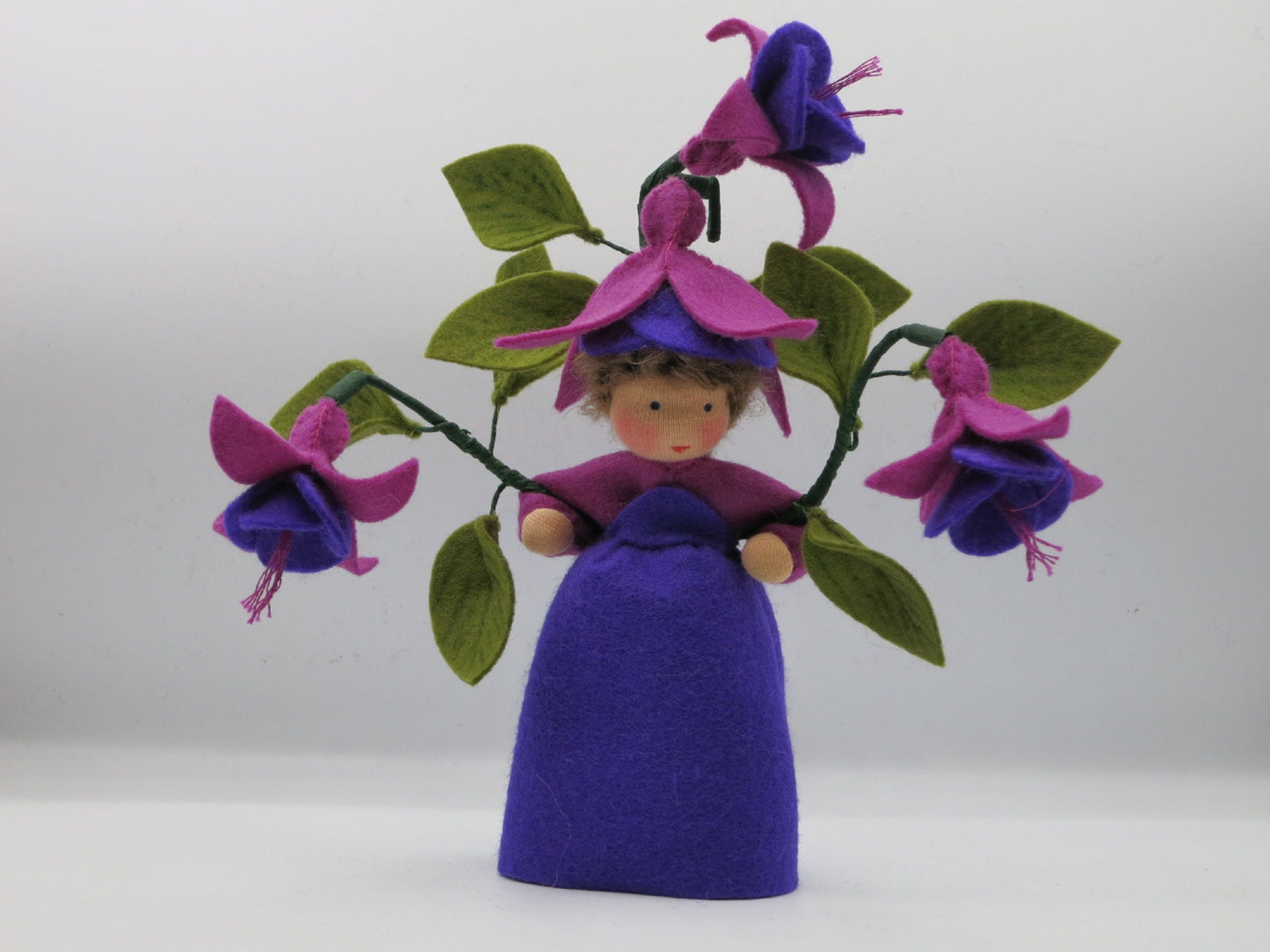 Handgefertigtes Fuchsien-Blumenkind aus Wollfilz mit violettem Kleid, Fuchsienblüten, grünen Blättern und Blütenhut.