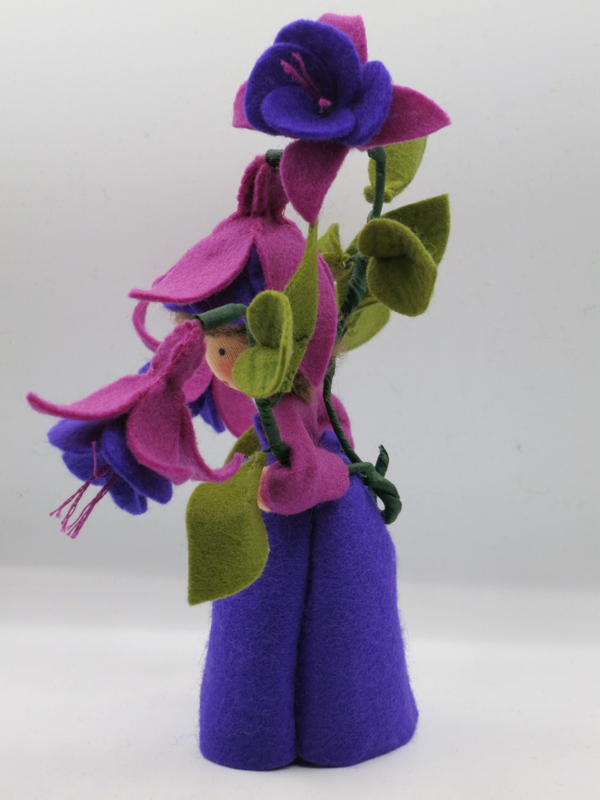 Rückansicht des Fuchsien-Blumenkinds aus Wollfilz mit violettem Kleid und hohem Stiel aus pink-violetten Blüten.