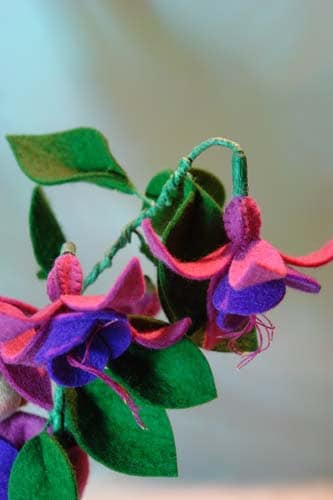 Detailansicht eines Zweigs mit handgefertigten Fuchsienblüten aus Wollfilz in Pink und Violett sowie grünen Blättern.