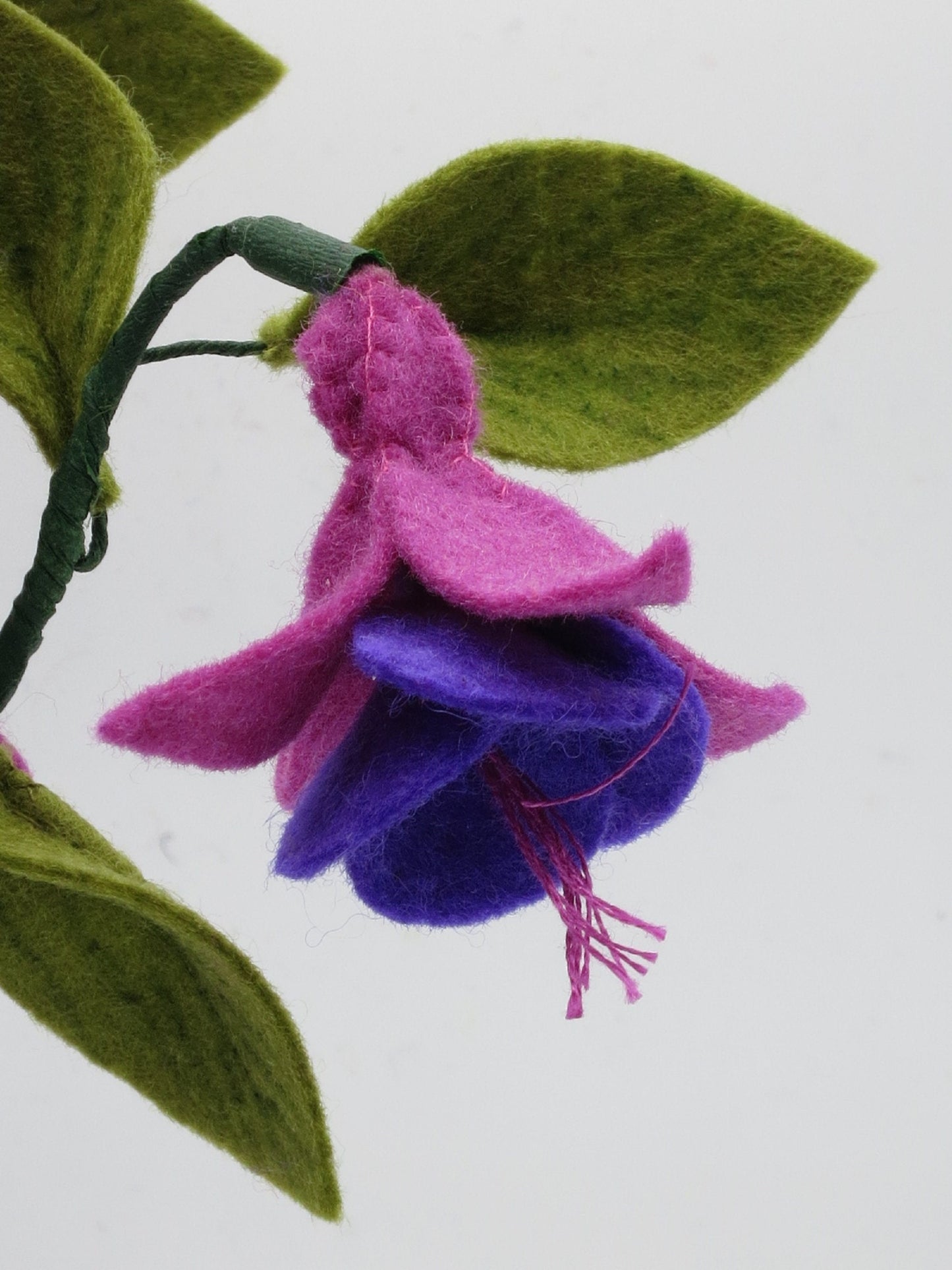 Detailansicht einer handgefertigten Fuchsienblüte aus Wollfilz in Pink und Violett mit grünen Blättern.