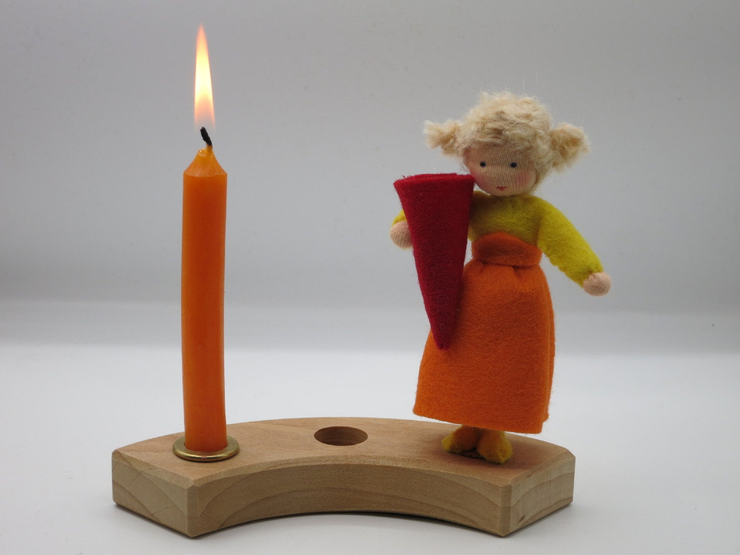 Geburtstagskind aus Wollfilz mit gelbem Oberteil, orangefarbenem Rock und roter Zuckertüte auf einem Holz-Kerzenring mit brennender Kerze.
