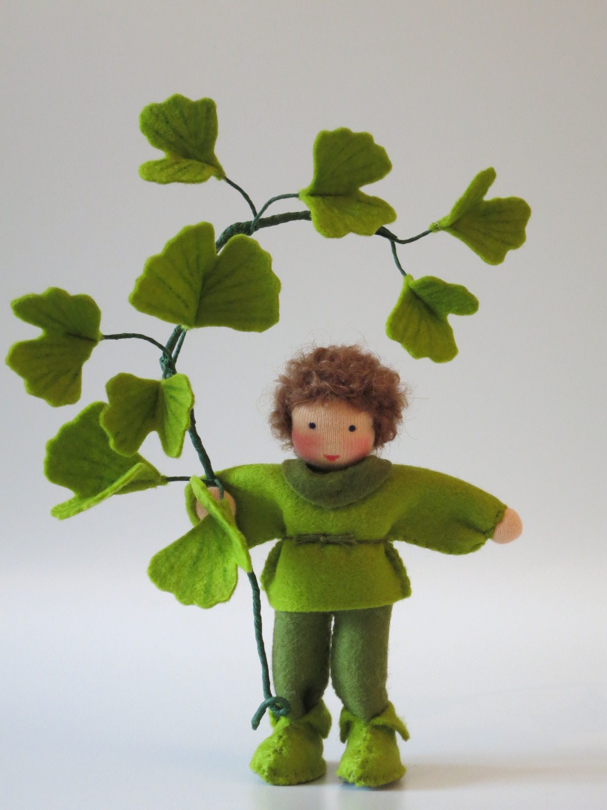 Frontalansicht des Ginkgo-Blumenkinds aus Wollfilz mit grüner Kleidung und einem Zweig mit großen Ginkgoblättern.