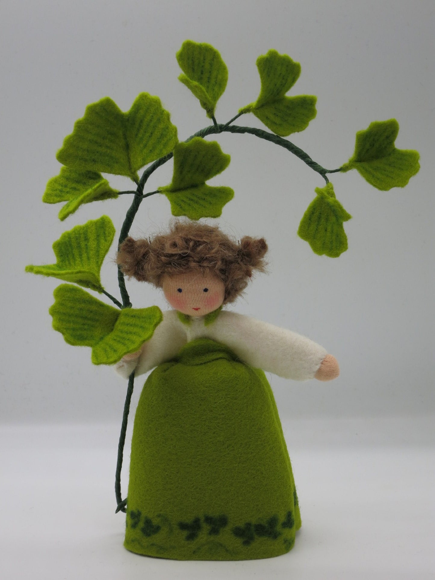 Frontalansicht des handgefertigten Ginkgo-Mädchens aus Wollfilz mit weißer Bluse, grünem Kleid und Ginkgozweig.