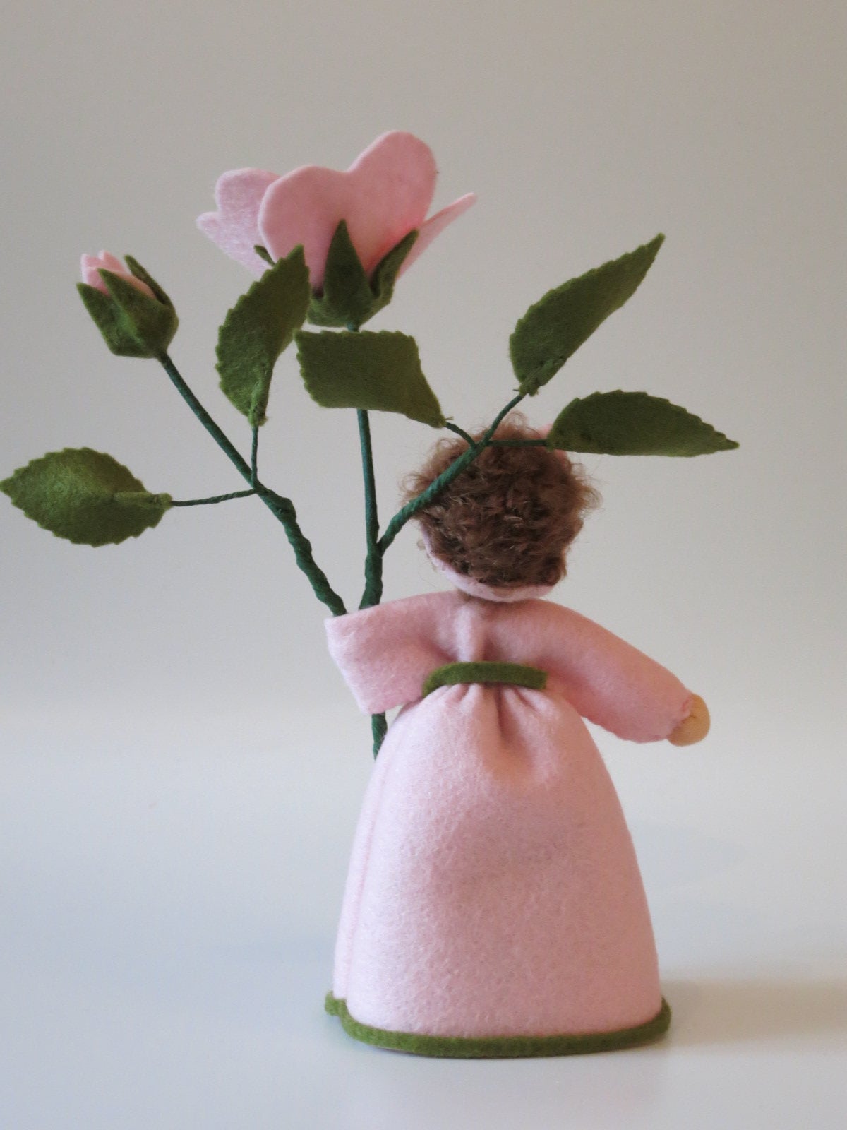 Rückansicht des Heckenröschen-Blumenkinds mit braunem Lockenhaar vor dem Blütenzweig, Wollfilz-Handarbeit.