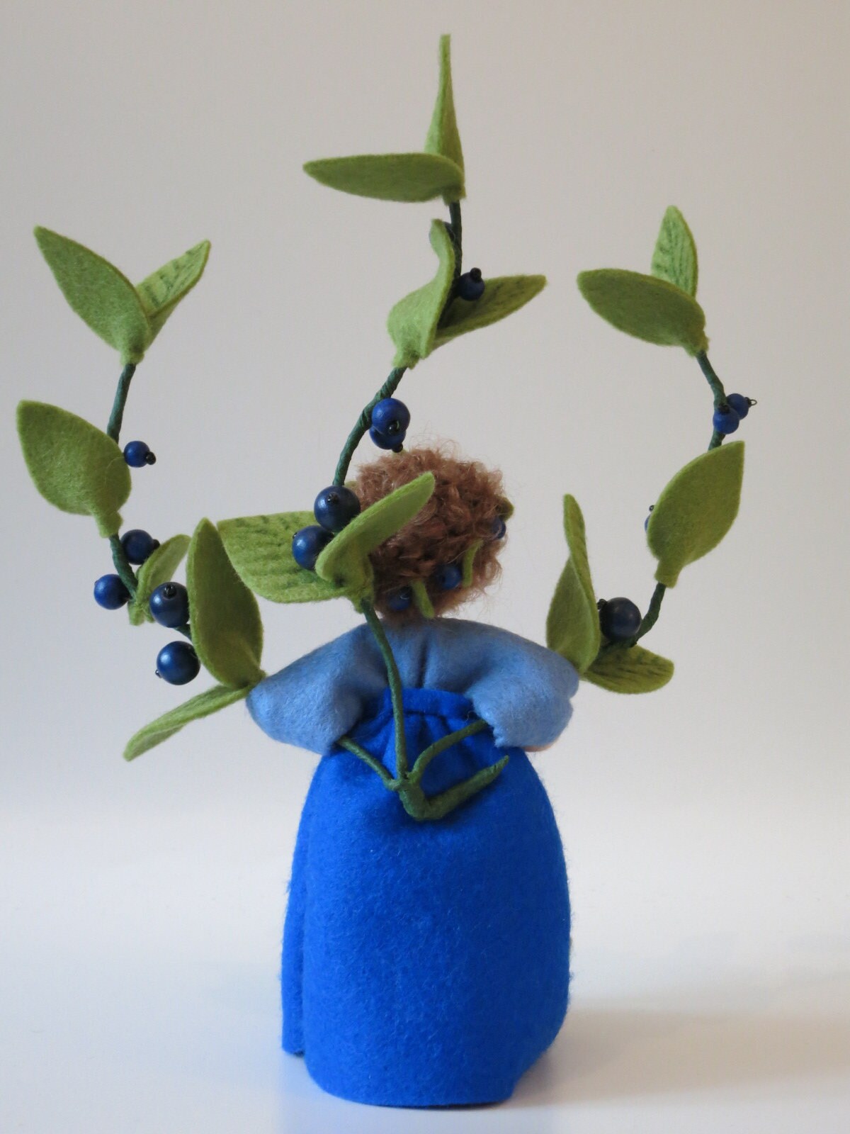 Rückansicht des Heidelbeere-Blumenkindes mit Beerenkranz im Lockenhaar und blauem Wollfilzkleid.