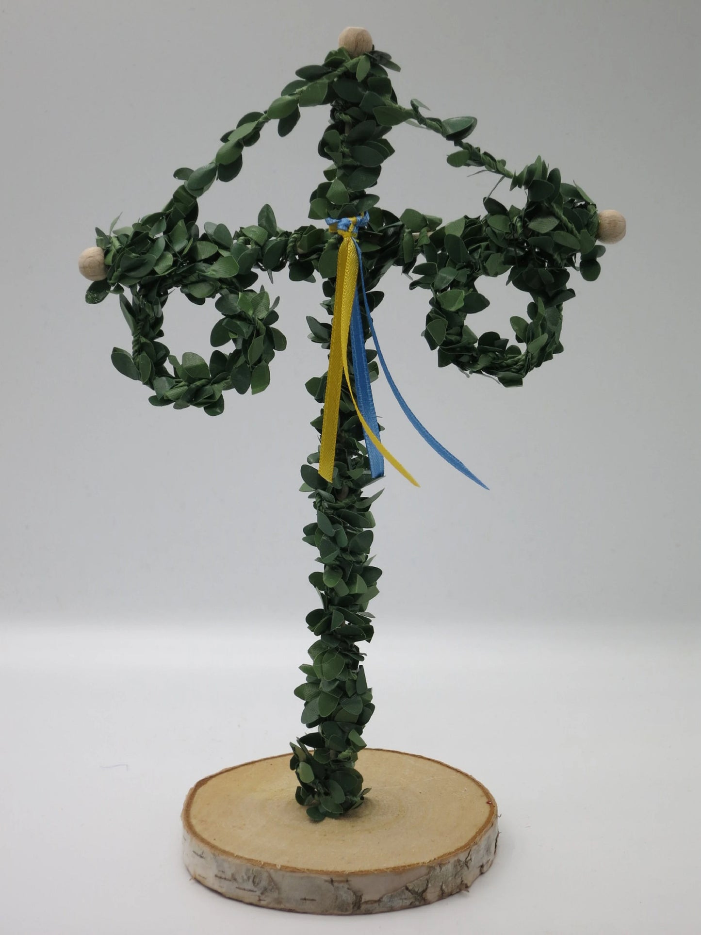 Mittsommerbaum-Variante mit grün umwickeltem Stab, zwei Laubkränzen und gelb-blauen Bändern auf Birkenholzscheibe.