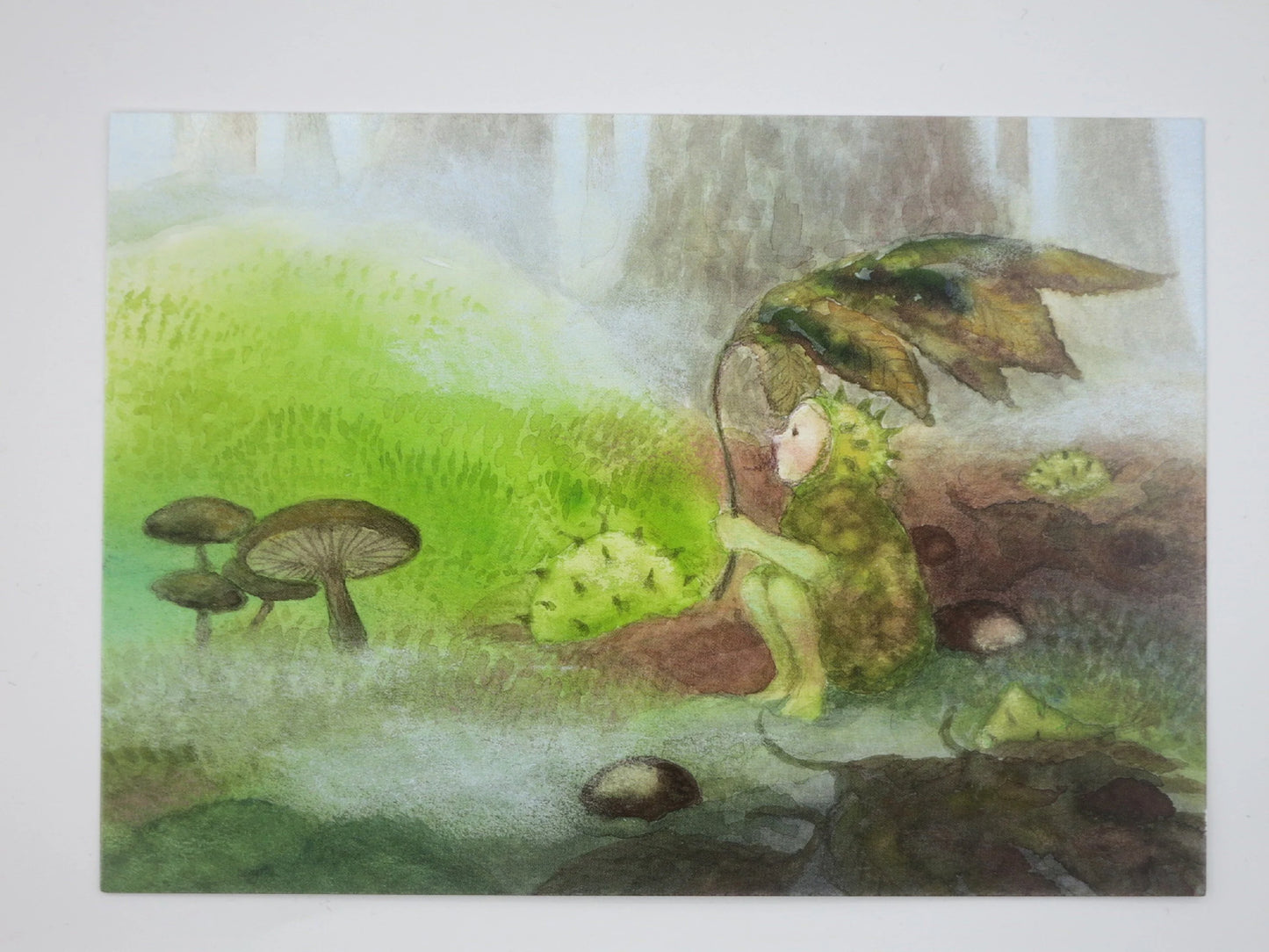 Postkarte Kastanienkind von Monika Speck: Naturgestalt aus Kastanienschale am moosigen Bachufer
