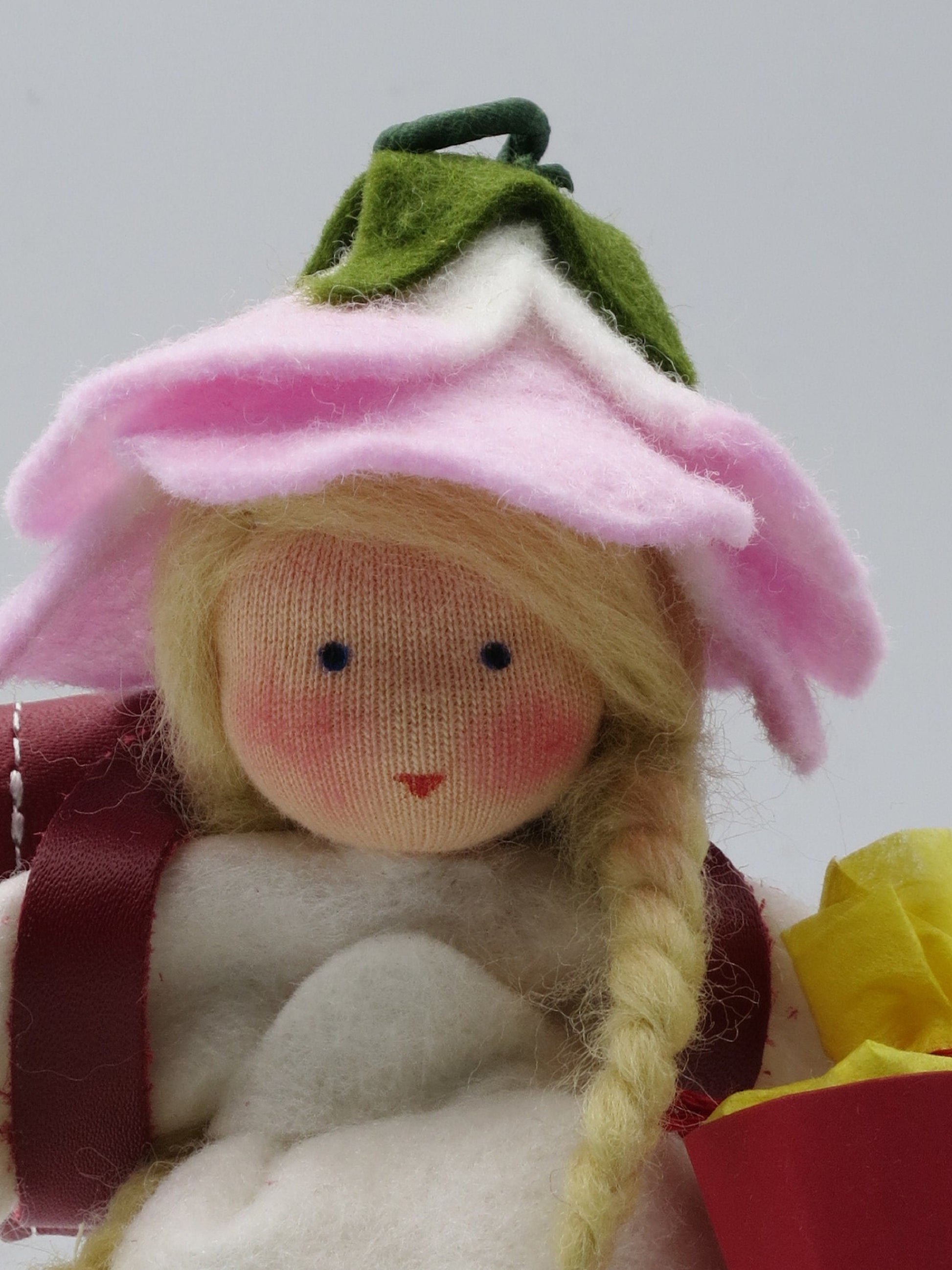 Gesichtsdetail des Schulmädchens mit rosa Rosenblütenhut und rotem Lederranzen aus Wollfilz.